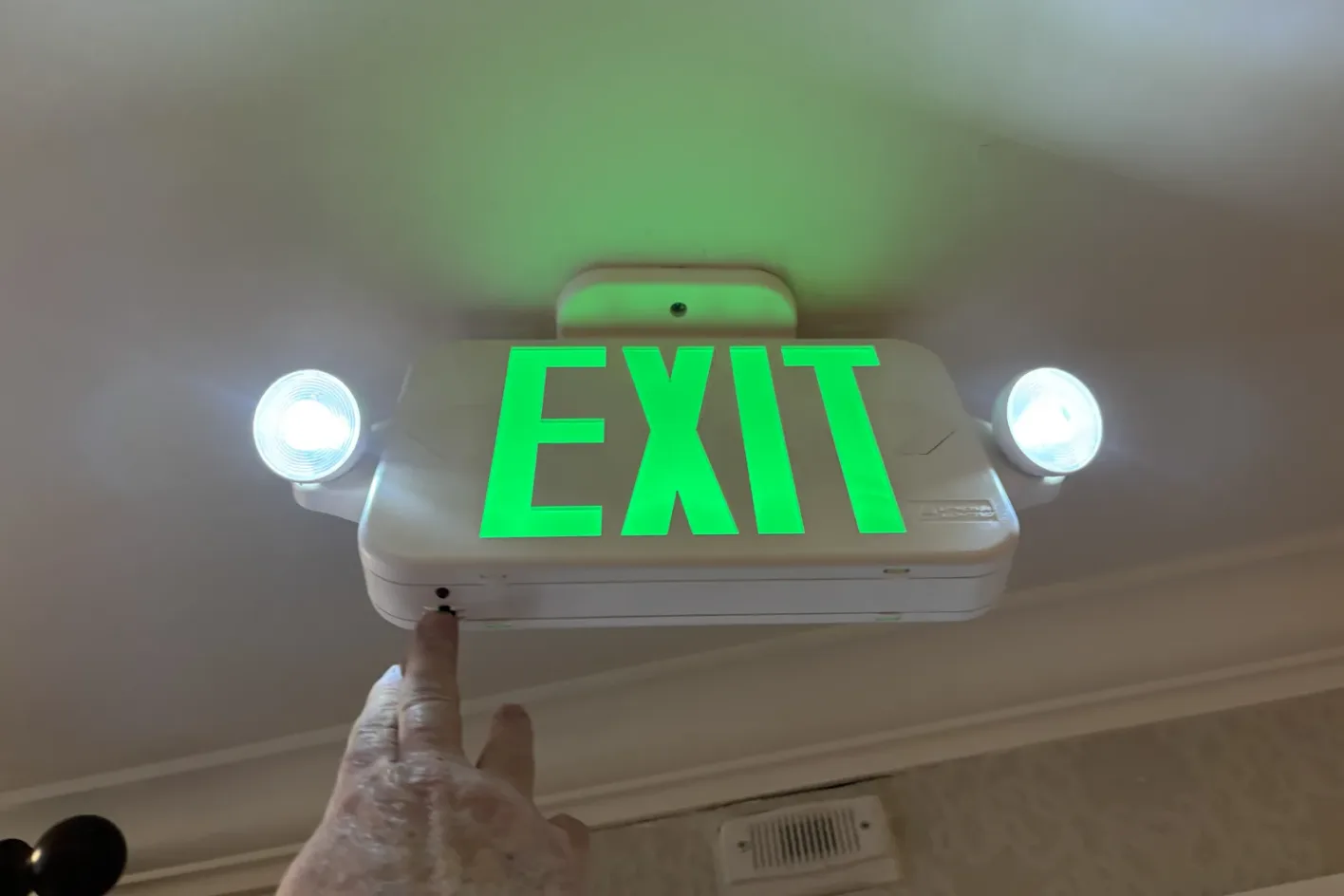 A hand pressing the test button on a green exit sign with emergency lamps lit during a monthly lighting check
